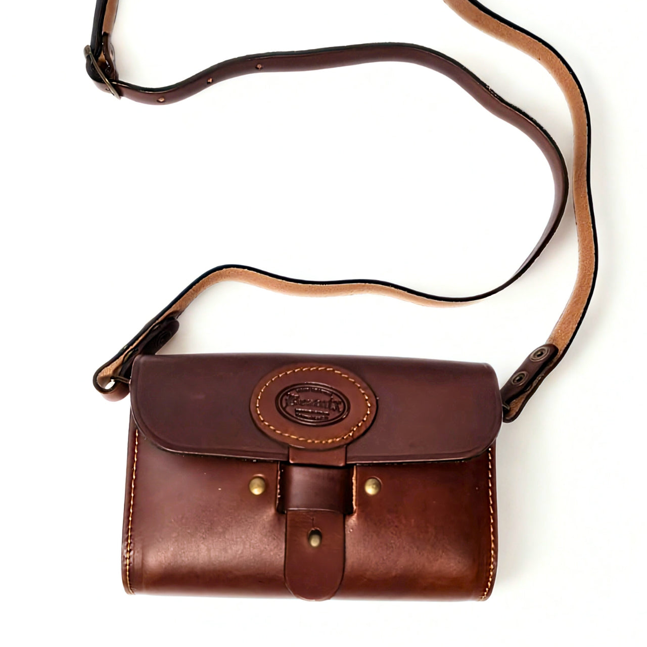 Beatnix Brown UZZI leather handbag, a rectangular brown leather bag with rounded edges, featuring a circular logo on the front flap that reads 'Beatnix Handbags.' The bag has visible stitching, two brass studs on the flap, and a leather strap with a brass buckle for closure. 3