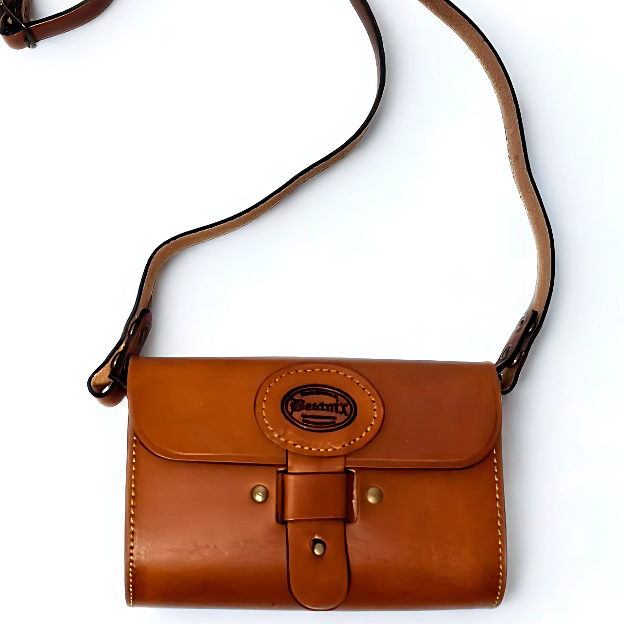 Beatnix tan UZZI leather handbag, a rectangular brown leather bag with rounded edges, featuring a circular logo on the front flap that reads 'Beatnix Handbags.' The bag has visible stitching, two brass studs on the flap, and a leather strap with a brass buckle for closure. 3